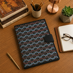 A patterned notebook or planner cover with a black leather tab closure sits on a wooden desk surrounded by books, a cup of pens, a small plant, glasses, and an open notebook.