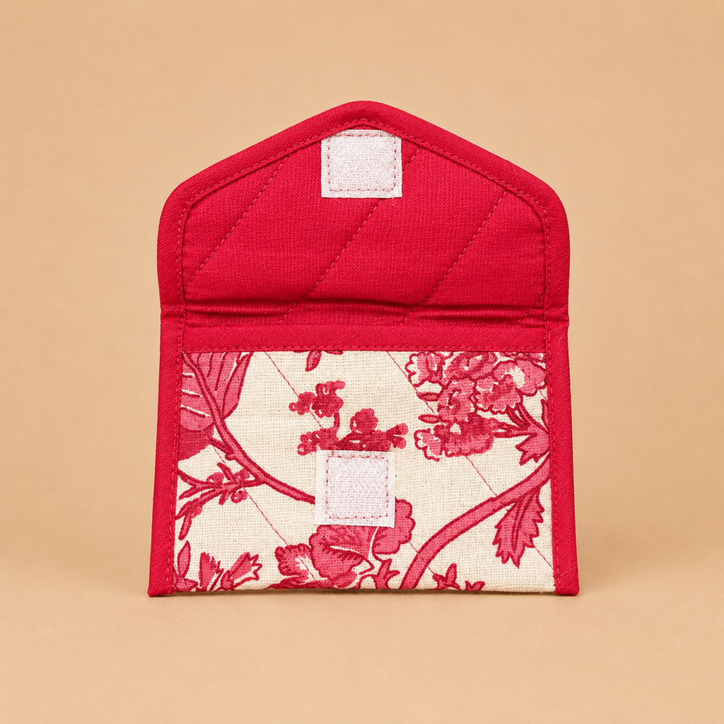 A small quilted fabric pouch in bright red, featuring a floral toile-style pattern panel in red and cream along the bottom half, with two small white label patches on the front, set against a solid beige background.