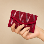 A hand holding a small envelope-style wallet made of deep red fabric with a gray and white ikat pattern, set against a solid beige background.