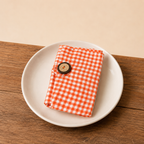 An orange and white gingham check fabric pouch with a brown button closure, resting on a white ceramic plate over a wooden tabletop.