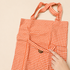 An orange and white gingham tote bag with a small brown button detail, shown with a hand pointing to the fabric texture against a light beige background.