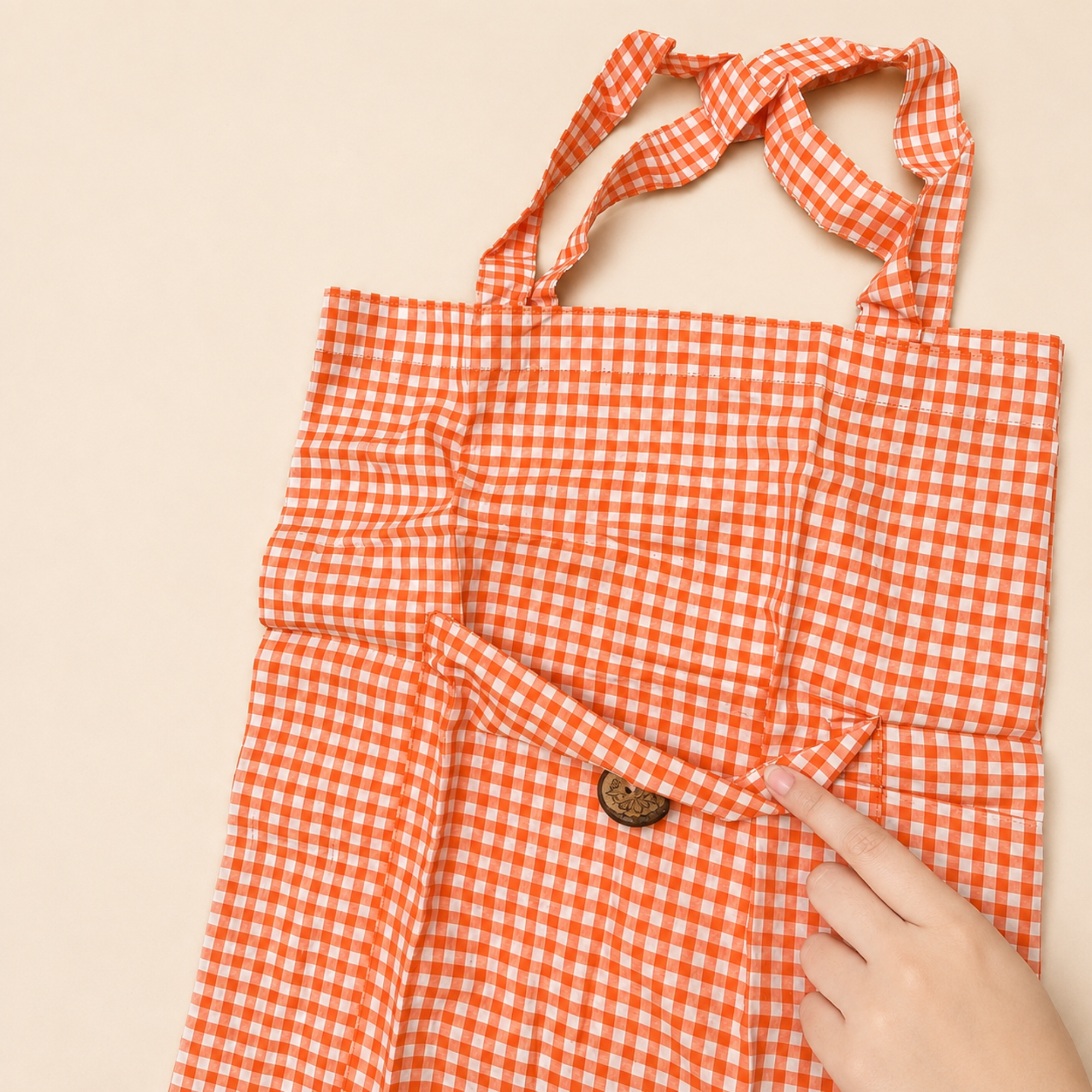 An orange and white gingham tote bag with a small brown button detail, shown with a hand pointing to the fabric texture against a light beige background.