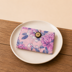 A pink floral fabric pouch with a button closure, displayed on a white ceramic plate over a wooden surface.