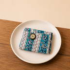 A small folded wallet or card holder with a teal, white, and gray geometric and floral pattern, featuring a round wooden button closure, displayed on a white ceramic plate over a wooden table.