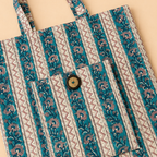 A fabric tote bag with a vertical striped pattern featuring teal blue, beige, and brown floral motifs, laid flat on a light beige background.