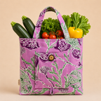 A purple fabric grocery tote bag with a green and white botanical pattern, filled with fresh vegetables including zucchini, tomatoes, lettuce, and a yellow bell pepper, set against a solid beige background.
