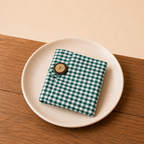 A small folded fabric item with a green and white gingham check pattern and a round wooden button, displayed on a white plate over a wooden surface.