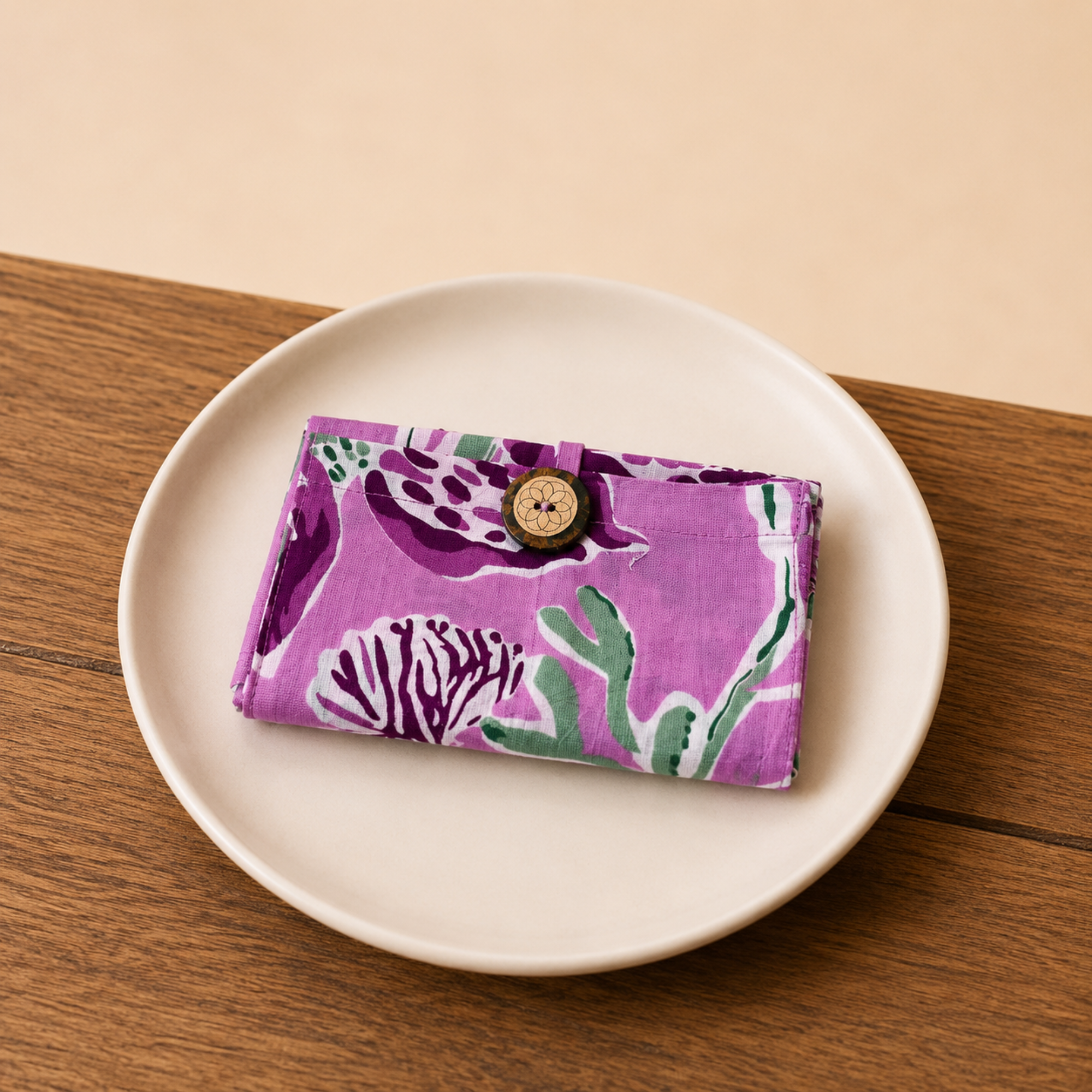 A small fabric pouch with a purple, green, and white botanical pattern and a wooden button closure, resting on a white ceramic plate over a wooden table.