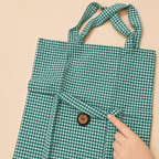 A green and white gingham tote bag with a small button detail, shown against a solid beige background with a hand pointing to the fabric texture.